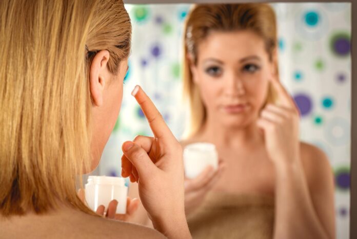 Beautiful young woman applying cream on her face in front a mirror in the bathroom at home.