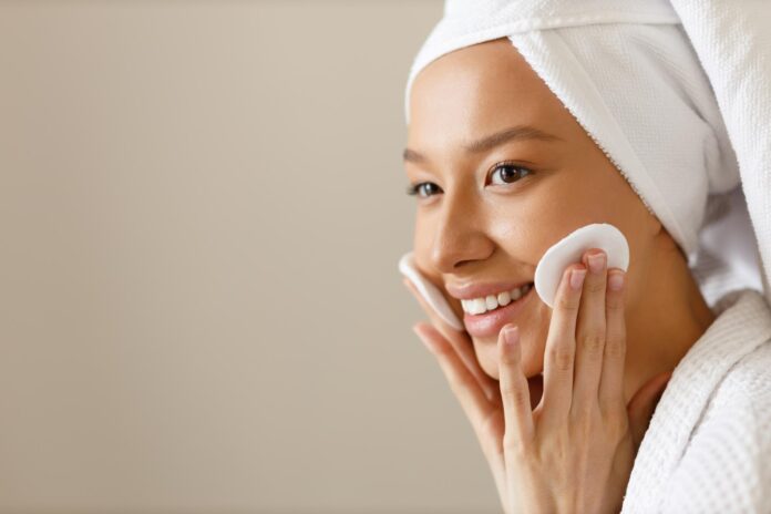 Pretty woman in robe and towel on her head wipes the skin of her face with cotton pads Cleansing and healthy body care after shower Closeup of girl on isolated background copy space